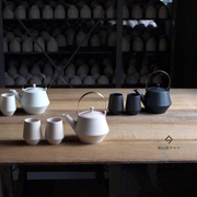 Yamatsu teapots and teacups arranged on a wooden table with shelving of unglazed pottery in background.

