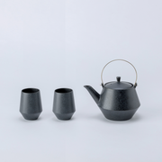 amatsu Frustum Japanese teapot with brass handle and two matching black teacups displayed on a white background.