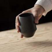 Hand holding a matte black Yamatsu Frustum teacup with a geometric shape and angled base, positioned over a light wooden surface.






