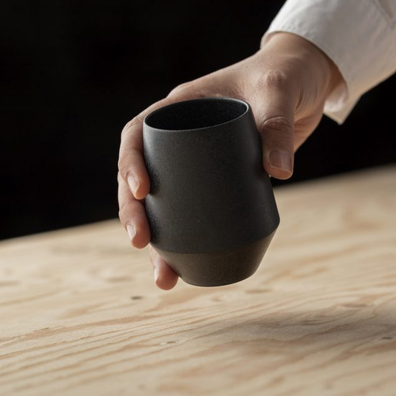 Hand holding a matte black Yamatsu Frustum teacup with a geometric shape and angled base, positioned over a light wooden surface.






