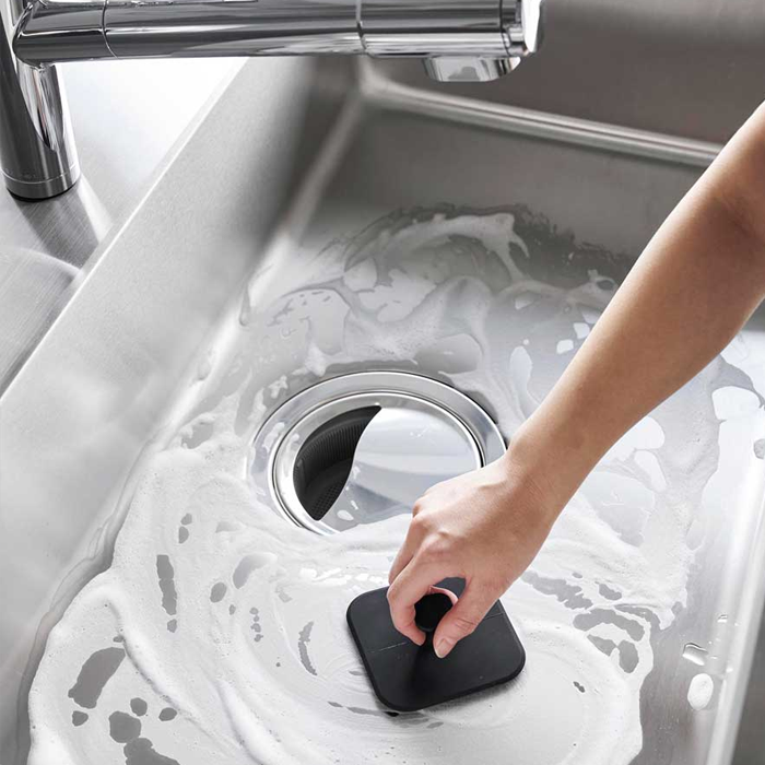 Person scrubbing a stainless steel sink with the black Yamazaki Tower Series silicone sponge creating soap suds.
