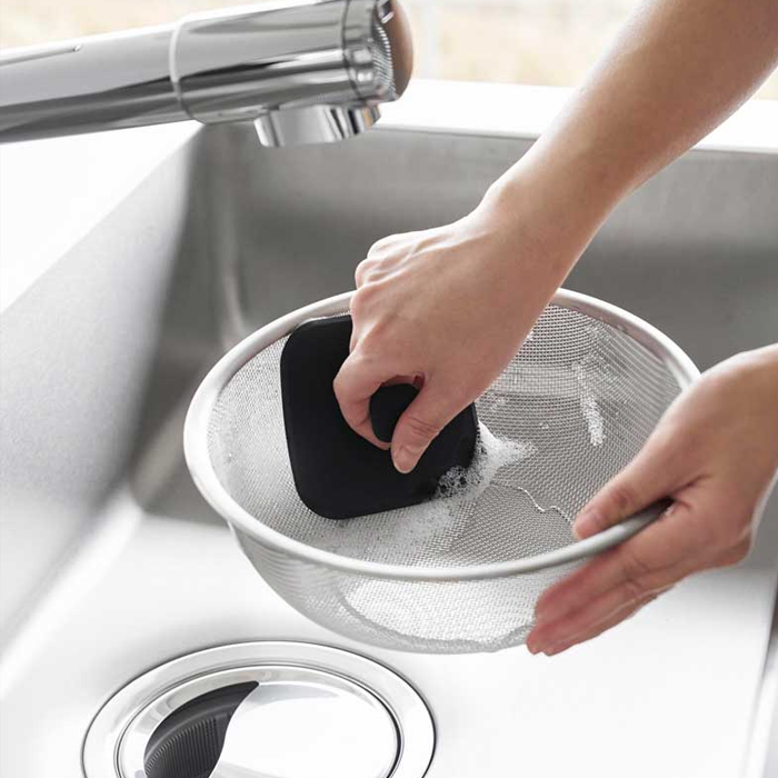A lifestyle image showing the Yamazaki Tower Series Silicone Sponge being used to easily clean a mesh strainer in a kitchen sink.

