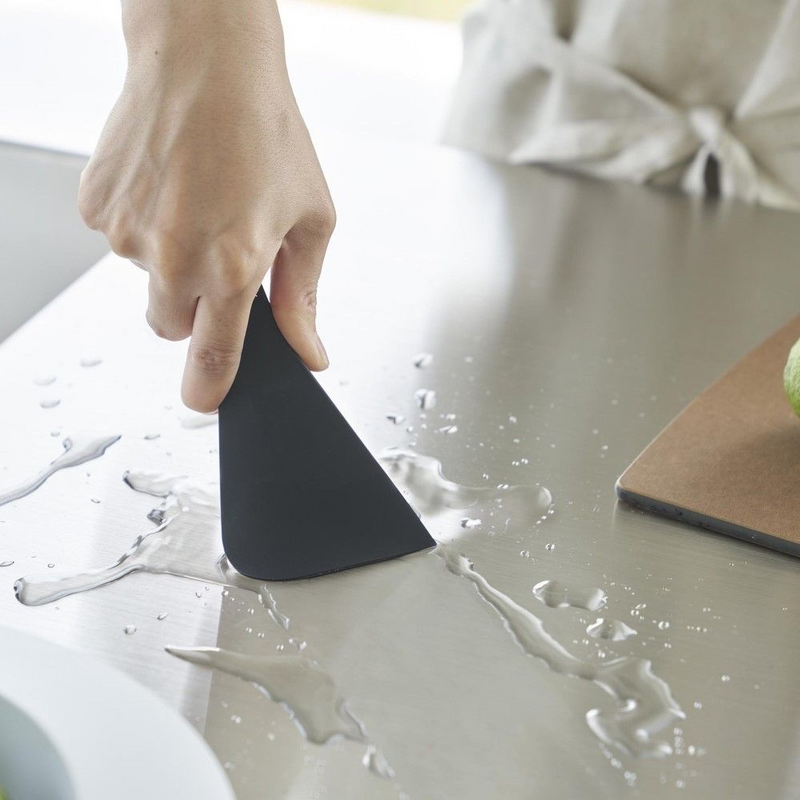 A hand using the Yamazaki Tower Series Silicone Cleaning Scraper to wipe up a water spill on a kitchen countertop, demonstrating its use.



