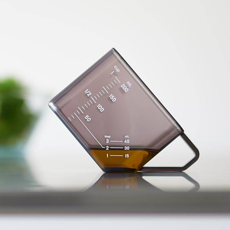 Yamazaki Tower Series Measuring Cup shown tilted to demonstrate tablespoon measurement mode, with clearly visible mL, cup, and tbsp scale markings on the dark smoke Tritan body.