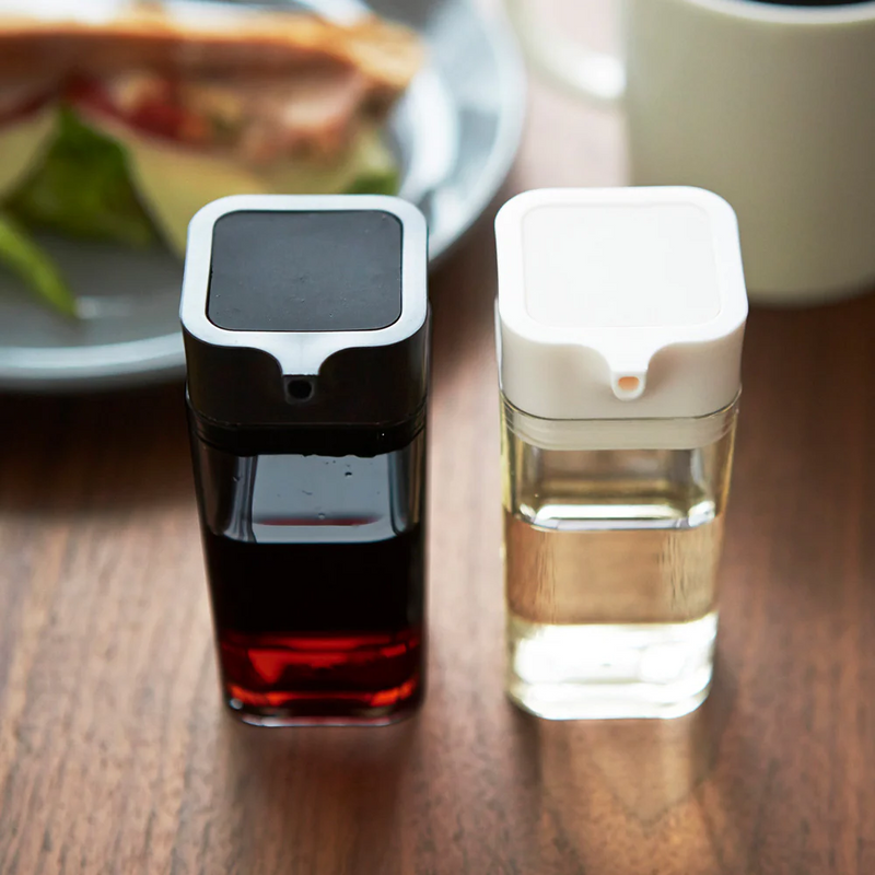 Yamazaki Tower Series One-Touch Soy Sauce Dispensers in black and white, with transparent glass bodies showing dark soy sauce and clear vinegar, on a wooden table