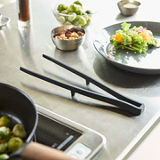 The Yamazaki Tower Series silicone serving tongs resting on their built-in stand on a stainless steel countertop, with a pan and plated salad in the background.