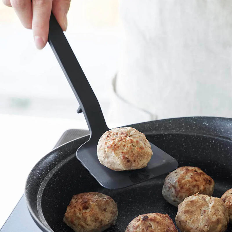 A lifestyle image of the Yamazaki Tower Series Small Silicone Turner easily lifting a meatball from a frying pan, showcasing its size and functionality.


