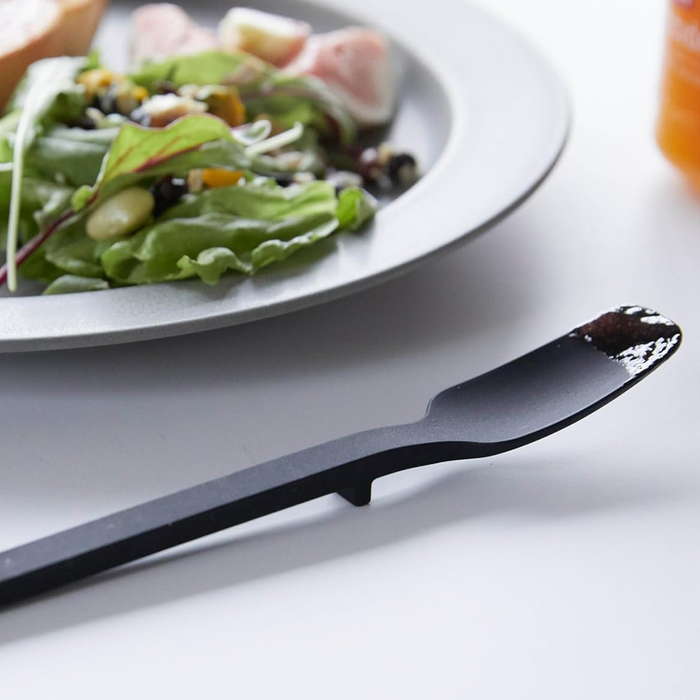 A Yamazaki Tower Series Silicone Scraping Spoon placed on a table beside a plate of salad, showing its raised tip design to keep surfaces clean.