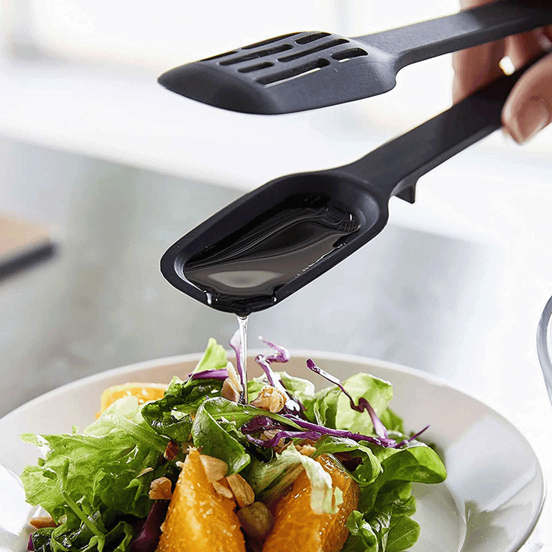 A pair of black Yamazaki Tower silicone tongs drizzling dressing from its spoon arm onto a salad.
