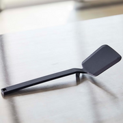 The Yamazaki Tower Series Silicone Turner in black, resting on a stainless steel countertop to demonstrate its integrated rest feature.
