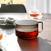 Yamazaki Tower Series low-profile glass teapot with matte black lid and geometric handle, filled with amber tea, shown on a wooden table with glass tea cups in the background.