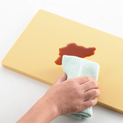 Hand using a white cloth to wipe up red sauce on a yellow cutting board.