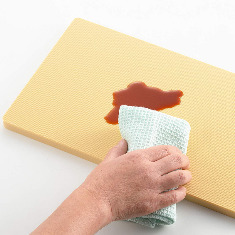 Hand using a white cloth to wipe up red sauce on a yellow cutting board.