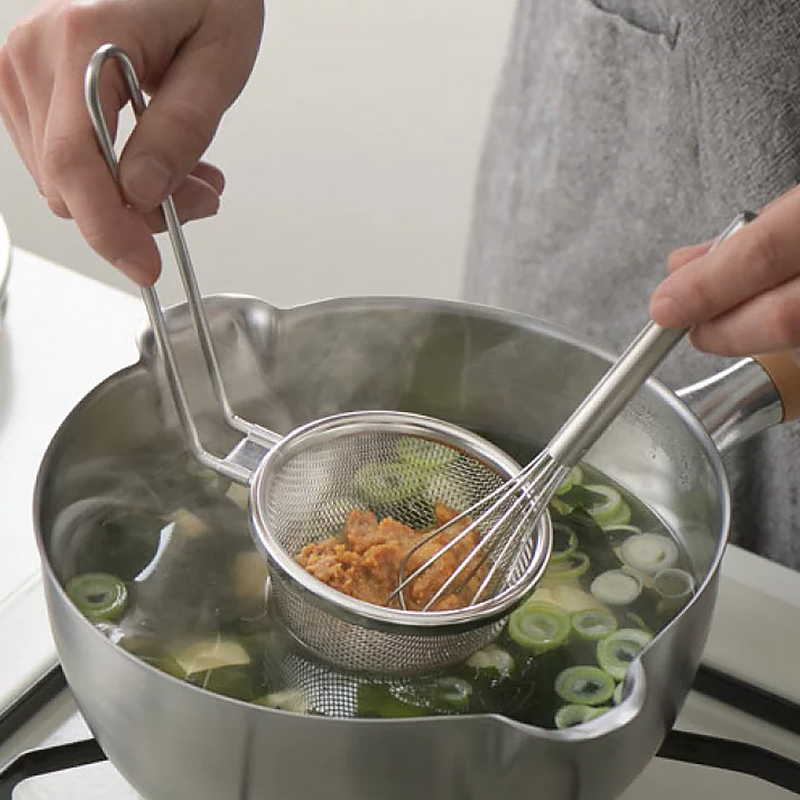 A person uses a Yoshikawa stainless steel strainer and a small whisk to dissolve miso paste into a pot of hot soup with green onions.