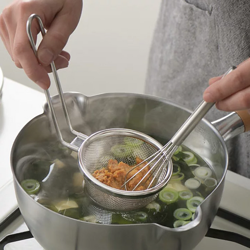 A person uses a Yoshikawa stainless steel strainer and a small whisk to dissolve miso paste into a pot of hot soup with green onions.