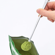 A hand holding a Yoshikawa long-handle spoon, scooping green powder from a foil bag.