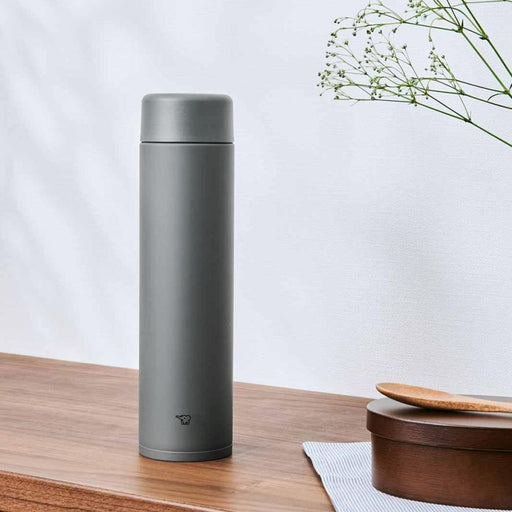 A matte grey Zojirushi SM-GA72 vacuum insulated flask standing on a wooden table next to a wooden container and spoon.