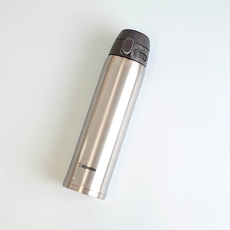 A silver Zojirushi SM-TA60-XA stainless steel vacuum insulated flask with a dark grey one-touch lid lying diagonally on a white background.