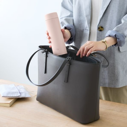 Person placing a pink Zojirushi flask into a grey handbag on a wooden table.

