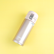 A champagne gold Zojirushi SM-KB36 vacuum insulated flask with a one-touch open lid lying diagonally on a yellow background.
