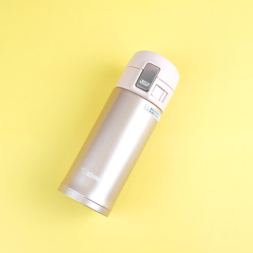 A champagne gold Zojirushi SM-KB36 vacuum insulated flask with a one-touch open lid lying diagonally on a yellow background.