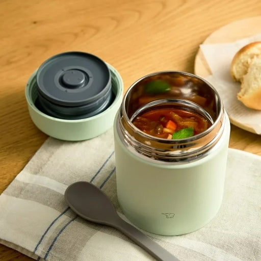 An open pale green Zojirushi SW-LA40 vacuum insulated food jar filled with vegetable soup, sitting on a striped napkin next to a grey spoon and the jar's lid.