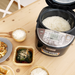 Open Zojirushi Micom Multifunctional Rice Cooker, model NL-GAQ18, displaying perfectly cooked rice inside, alongside dishes of various prepared foods on a wooden table.