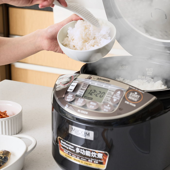 Person scooping freshly cooked white rice from the Zojirushi Micom NL-GAQ10 Multifunctional Rice Cooker. The cooker's digital display is visible, showing the current settings, and a couple of side dishes are placed beside it on the countertop.
