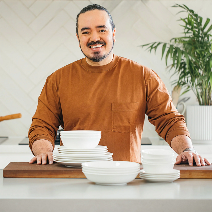 Adam Liaw standing behind his Everyday Noritake 12-Piece Dinnerware Set, featuring plates and bowls arranged on a kitchen countertop.
