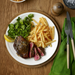 A steak meal with fries and greens served on the Adam Liaw Everyday Noritake 25cm large plate, alongside cutlery and a drink.