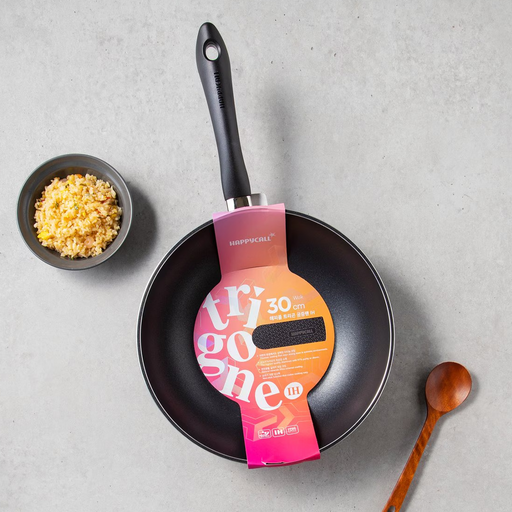 Frying pan with a pink packaging label on a gray surface, surrounded by a bowl of garlic and a wooden spoon.