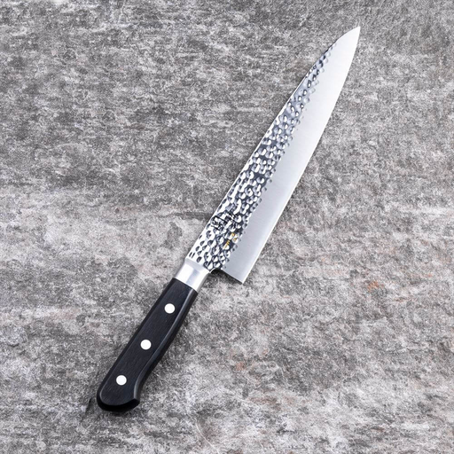 The Kai Seki Magoroku Hammered Chef Knife (210mm) laid on a grey stone surface, showcasing the beautiful tsuchime (hammered) finish on the blade and its ergonomic black handle.
