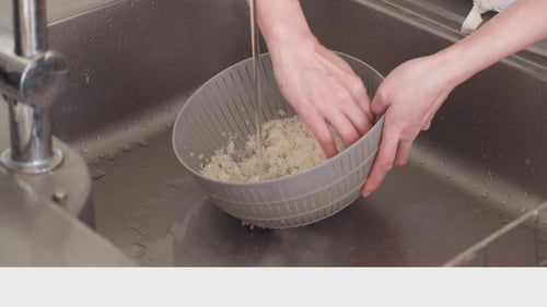 A person using their hands to wash rice in the grey Like-it colander under running water in a sink.