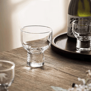 A lifestyle setting featuring the Toyo Sasaki sake glass on a wooden table, with a sake bottle and another glass in the background.


