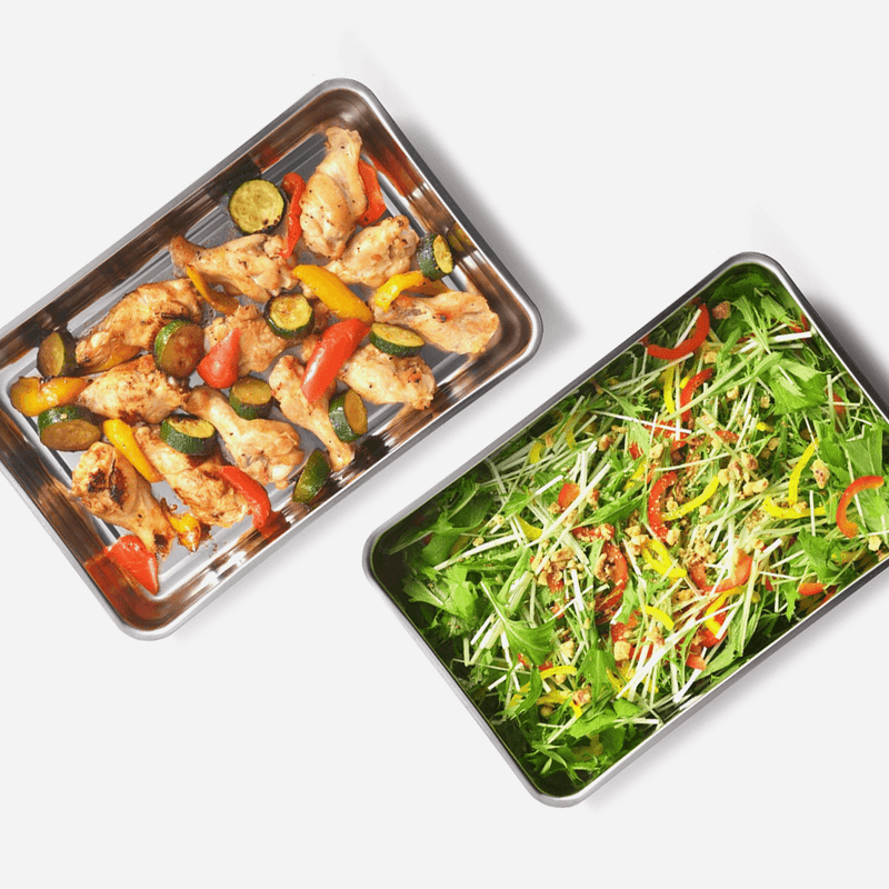Two Yoshikawa stainless steel containers used for serving, one filled with roasted chicken wings and vegetables, the other with a fresh salad.
