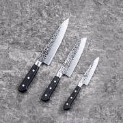 The Kai Seki Magoroku hammered 3-piece knife set, including a santoku knife, a chef knife, and a utility knife, all with textured blades and black handles, arranged on a surface.
