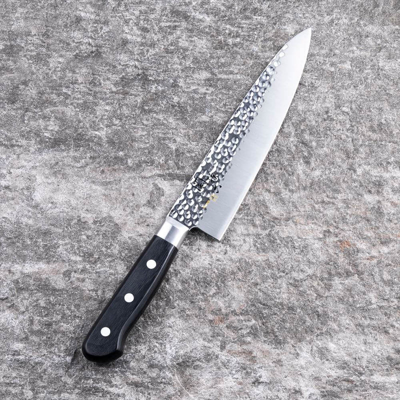The Kai Seki Magoroku Hammered Chef Knife (180mm) laid on a grey stone surface, showcasing the beautiful tsuchime (hammered) finish on the blade and its ergonomic black handle.
