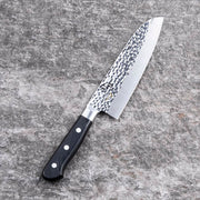 The Kai Seki Magoroku Hammered Santoku Knife (165mm) laid on a grey stone surface, showcasing the beautiful tsuchime (hammered) finish and its classic Santoku blade shape.
