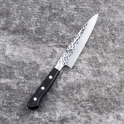 The Kai Seki Magoroku Hammered Utility Knife (120mm) laid on a grey stone surface, showcasing the tsuchime (hammered) finish on its slender blade.
